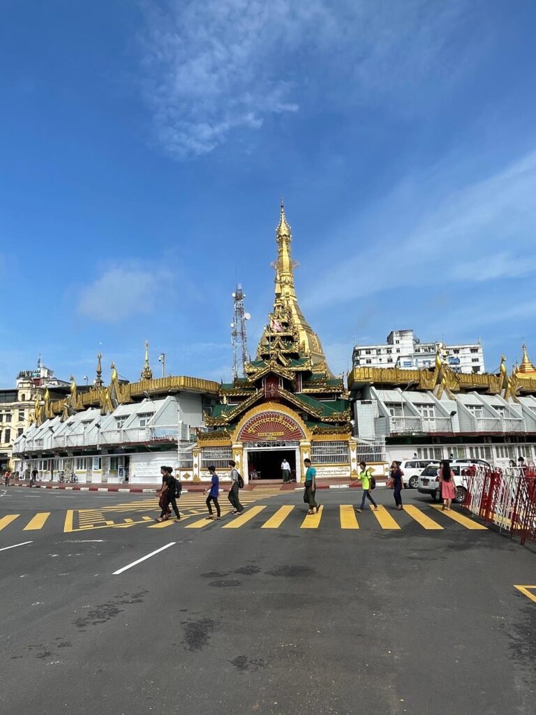 Sule Pagoda in Yangon. It is a white and green building with a golden pagoda pointing up from the centre. To the right is a red and white barricade which signifies a government building or other important structure in Myanmar.