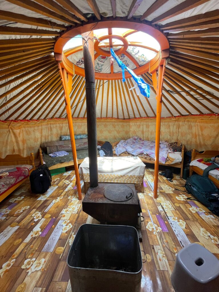 An accommodation ger in Mongolia. There are four beds around the outside (plus one not in this image). An iron stove sits in the centre of the room, whilst a bin and small stool are at the bottom of the image