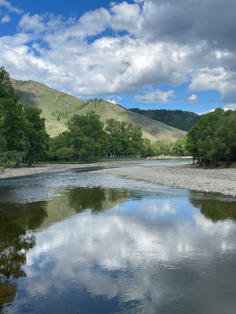 A river in front of green/brown hills in the Mongolian countryside during summer