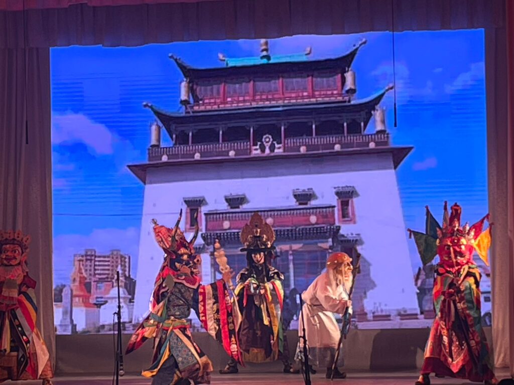 A Tibetan Buddhist Tsam dance, where performers are wearing colourful masks in front of an image of a Buddhist temple