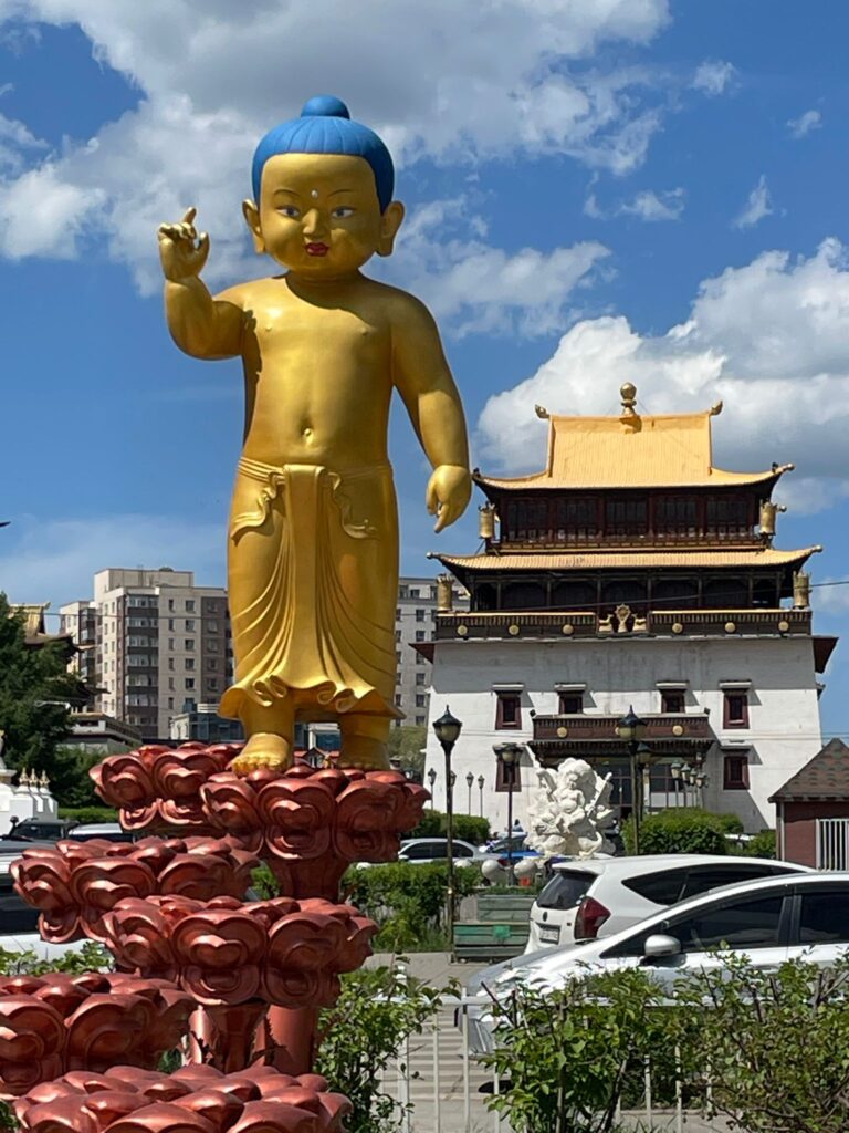 A golden statue of Buddha outside Gandantegchinlen Monastery in Ulaanbaatar, Mongolia. You can see a Buddhist temple in the background.