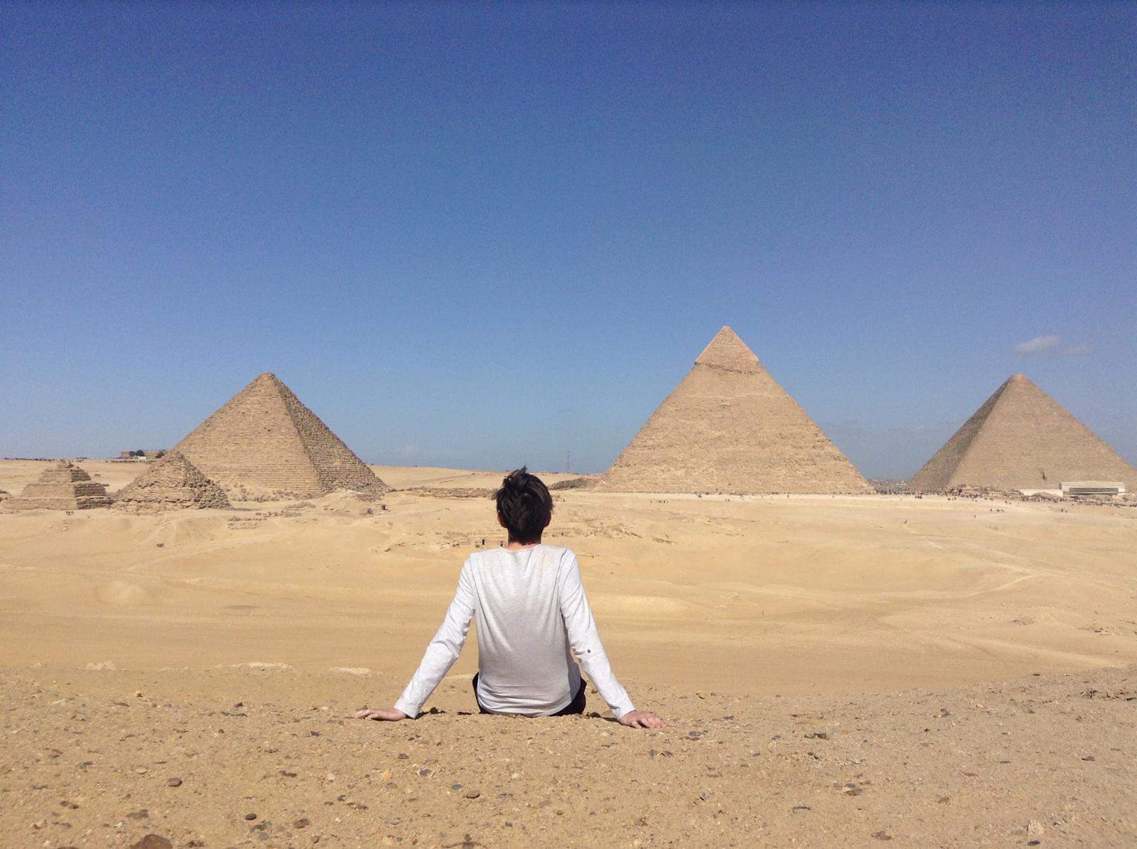 An image of Alex sat in the sand with his back to the camera, whilst looking at the Great Pyramids of Giza which stand tall in the background