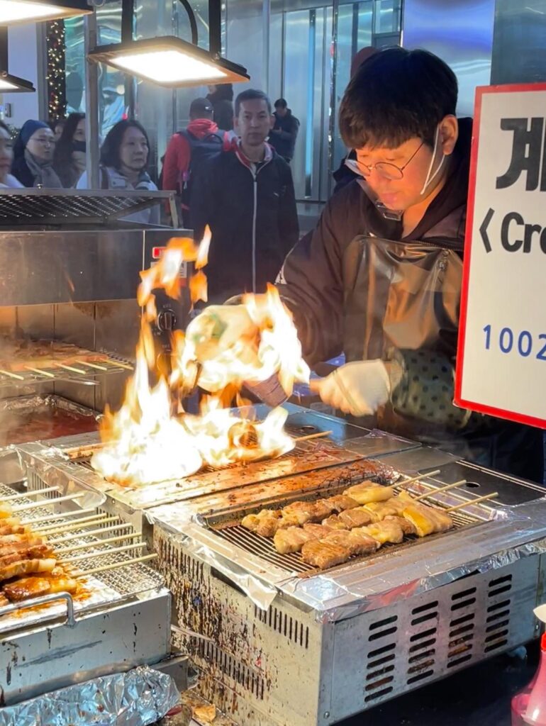 A vendor at Myeongdong night market using flames to cook chicken for skewers