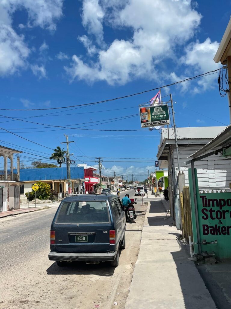 A street in Corozal, Belize. Buildings are small and similar to shacks. There are several cars and motorbikes on the road