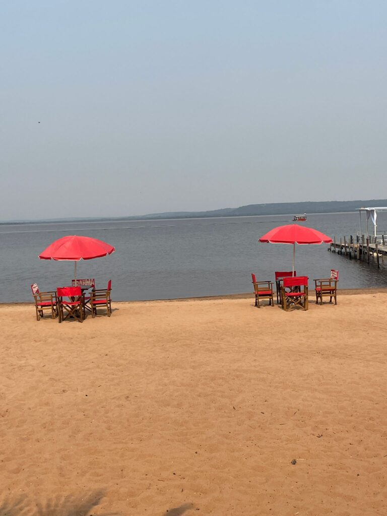 Two tables, each surrounded by four chairs with a red parasol in the middle. They are located on the yellow sands of San Bernardino, Paraguay, with Lake Ypacarai visible in the background