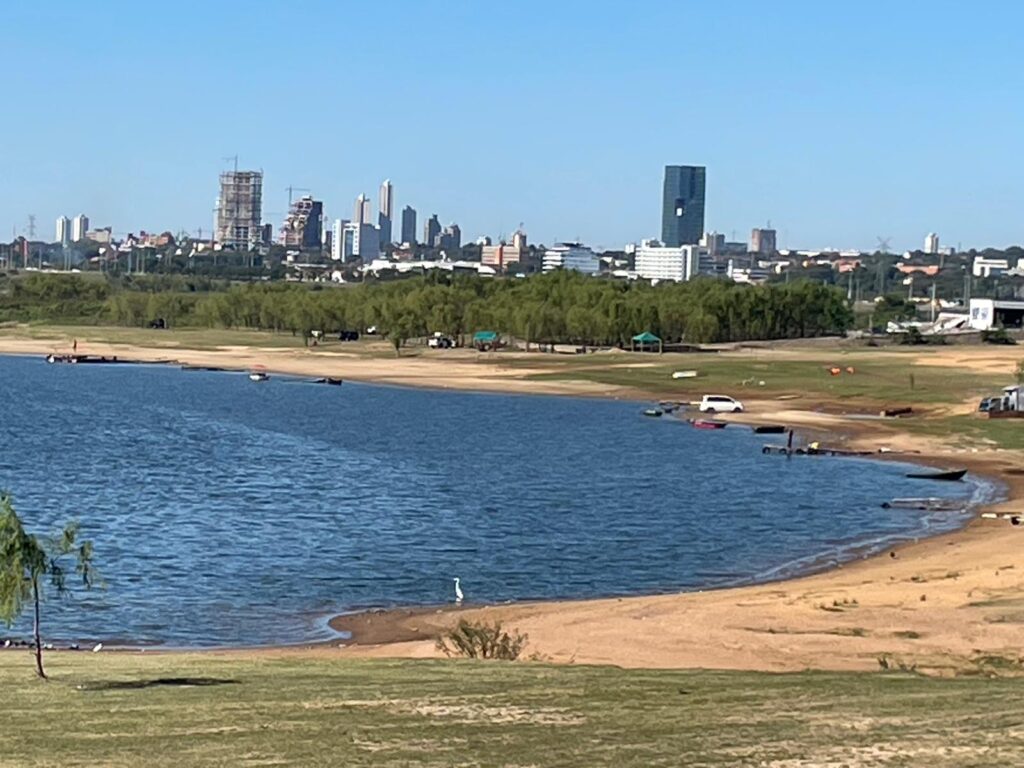 The blue water of the Paraguay River and yellow sandy beaches with skyscrapers visible in the distance. This is La Costanera in Asuncion