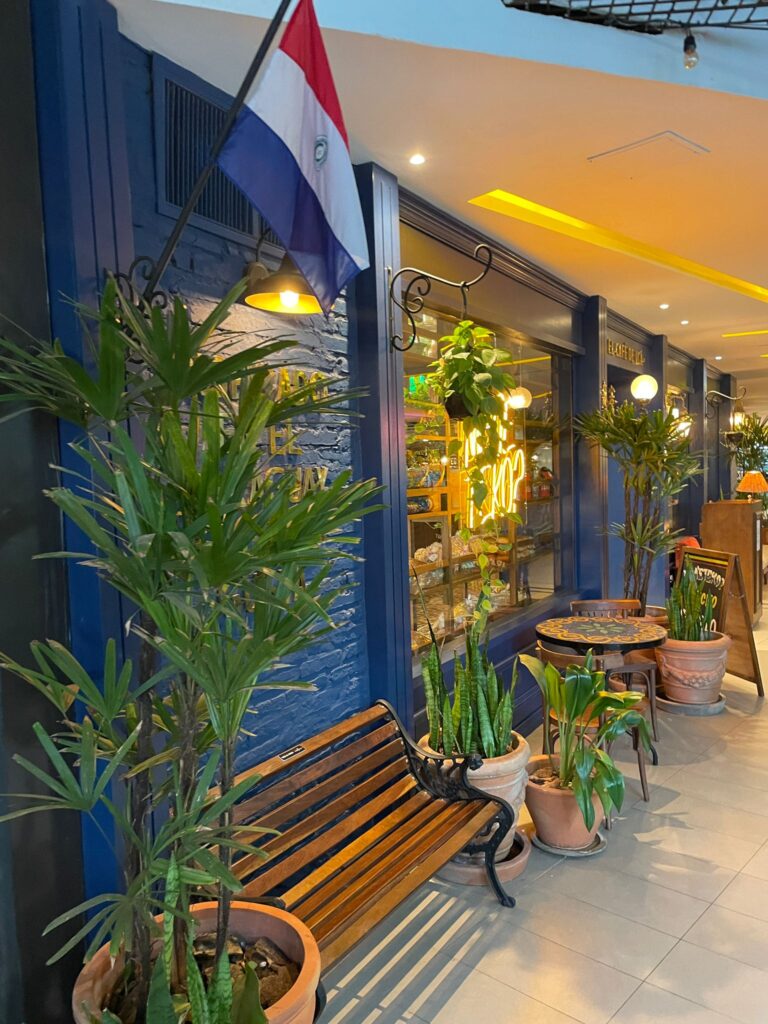 A restaurant with blue walls and the Paraguay flag outside, just above a bench. It is located in Asuncion's Paseo la Galeria