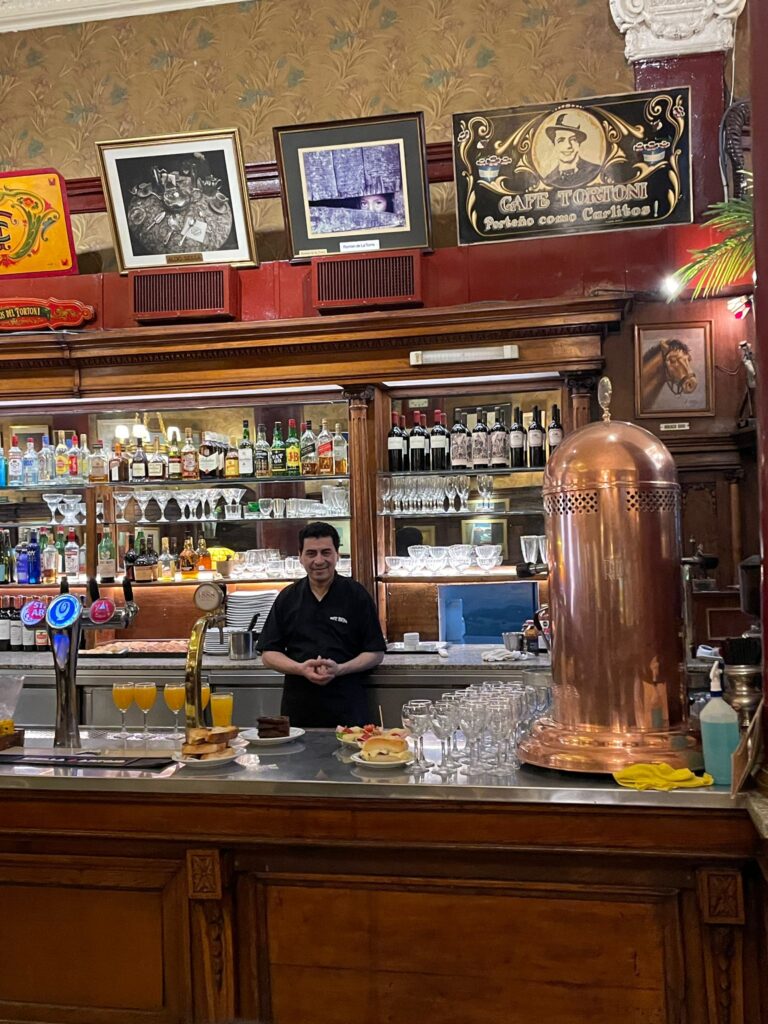 A bartender stands proudly at Cafe Tortoni in Buenos Aires, with a series of juices, cakes and sandwiches in front of him. There are several paintings on the wall above him