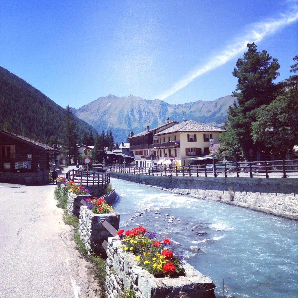 A bright blue river flowing through a village (Champoluc in Italy), with mountains in the background and colourful flowers to the left