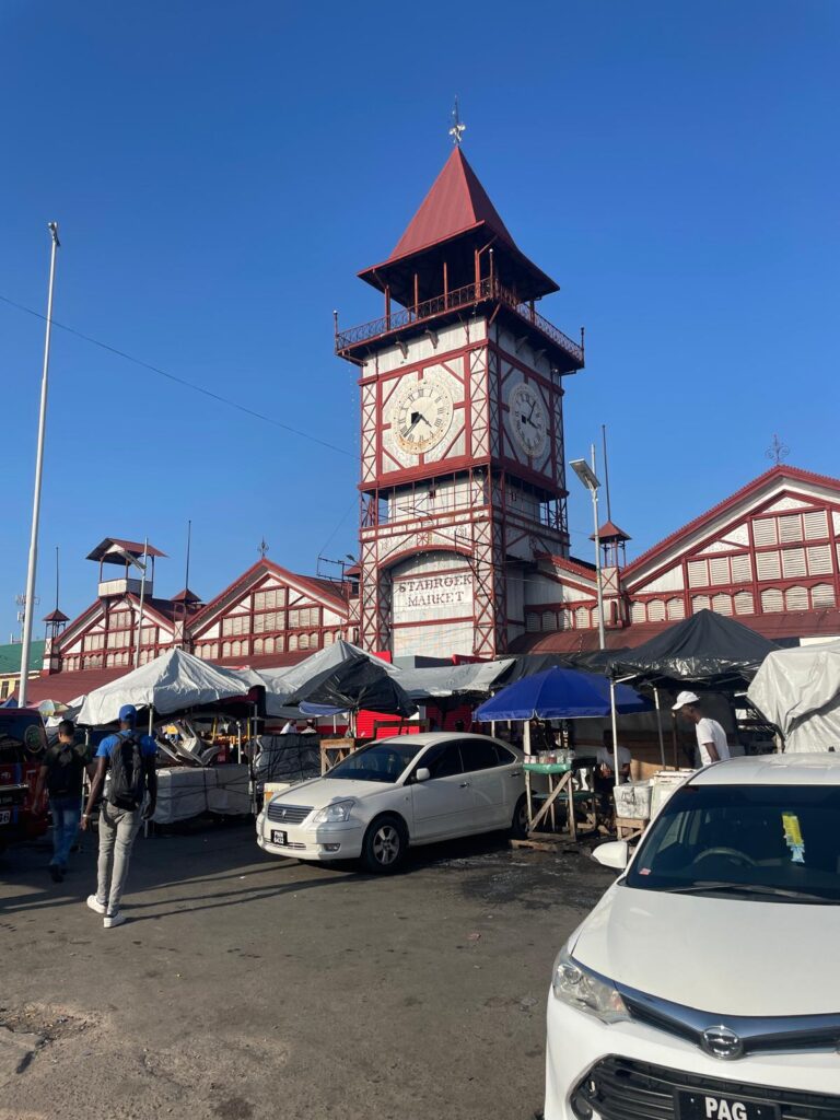 The famous clocktower outside Stabroek Market in Georgetown, Guyana. The tower is painted red and white with the name of the market below the clock. In front of the tower are several market stalls with locals selling a variety of goods