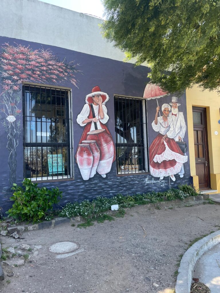 An image painted on a building in Colonia del Sacramento, where a man bangs drums whilst a couple shelter under an umbrella. All three characters are wearing red and white.