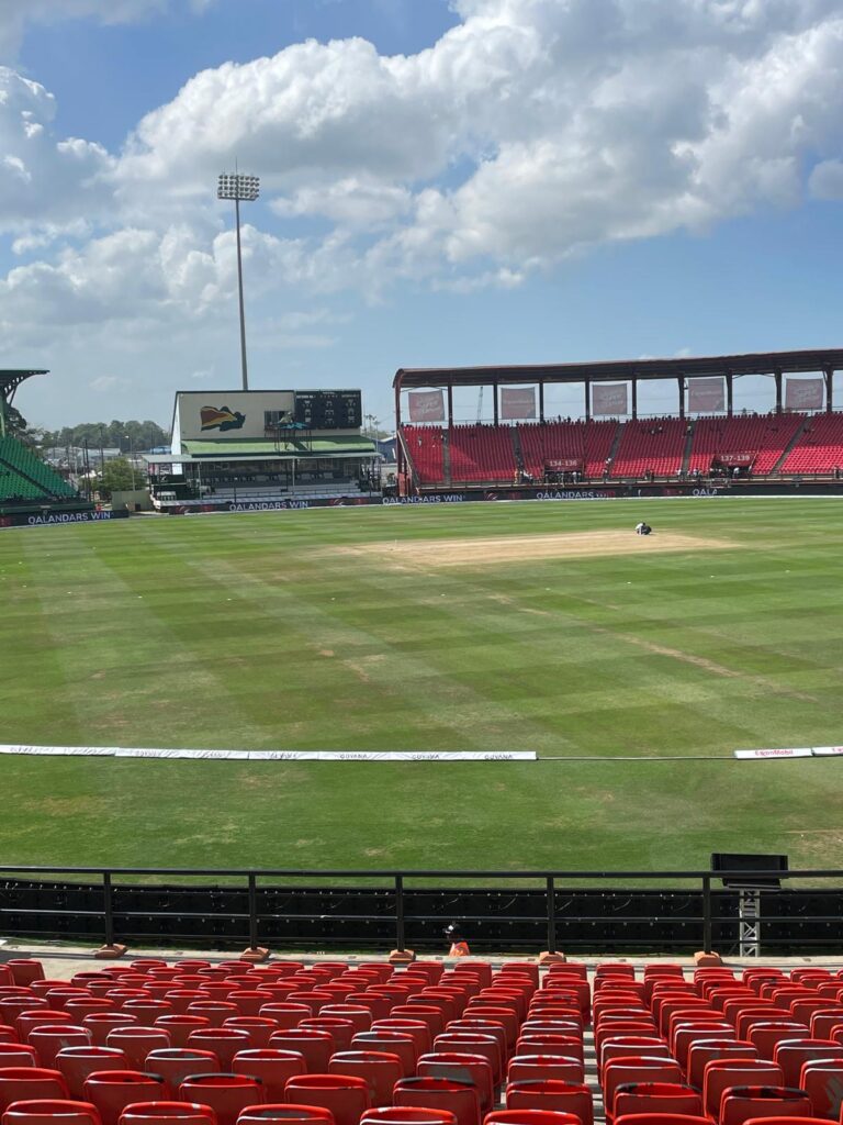 An image of Providence Stadium in Georgetown, Guyana. You can see the cricket pitch with red and green stands in the background. In the middle is a much smaller stand with Guyana's flag displayed