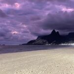 Purple skies over Rio de Janeiro's famous Ipanema Beach. The yellow sand is covered in footprints as people leave due to the sun setting. You can see the bright lights of several skyscrapers in the distance.