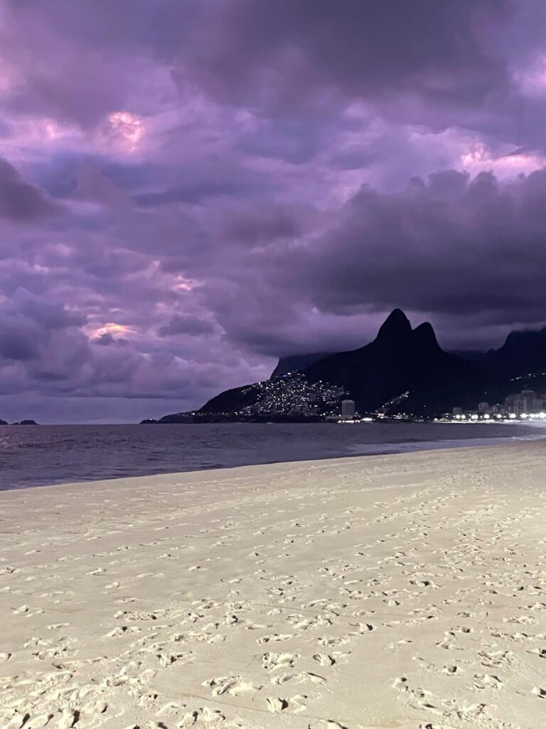 Purple skies over Rio de Janeiro's famous Ipanema Beach. The yellow sand is covered in footprints as people leave due to the sun setting. You can see the bright lights of several skyscrapers in the distance.