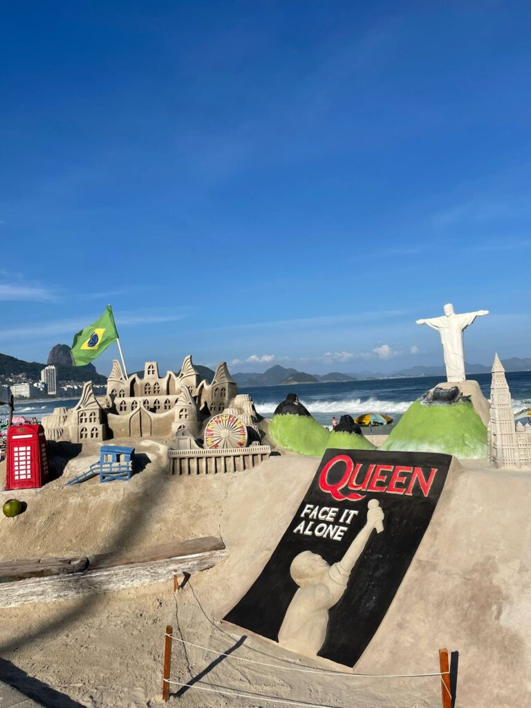 A sand display on Copacabana Beach in Rio de Janeiro. It shows some of the mountains in Rio with Christ the Redeemer on top. There is also a sign that says "Queen, face it alone".